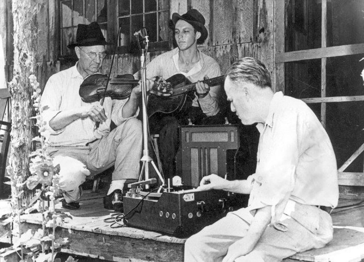 Vance Randolph at a field recorder on a porch while a fiddler and a guitarist play